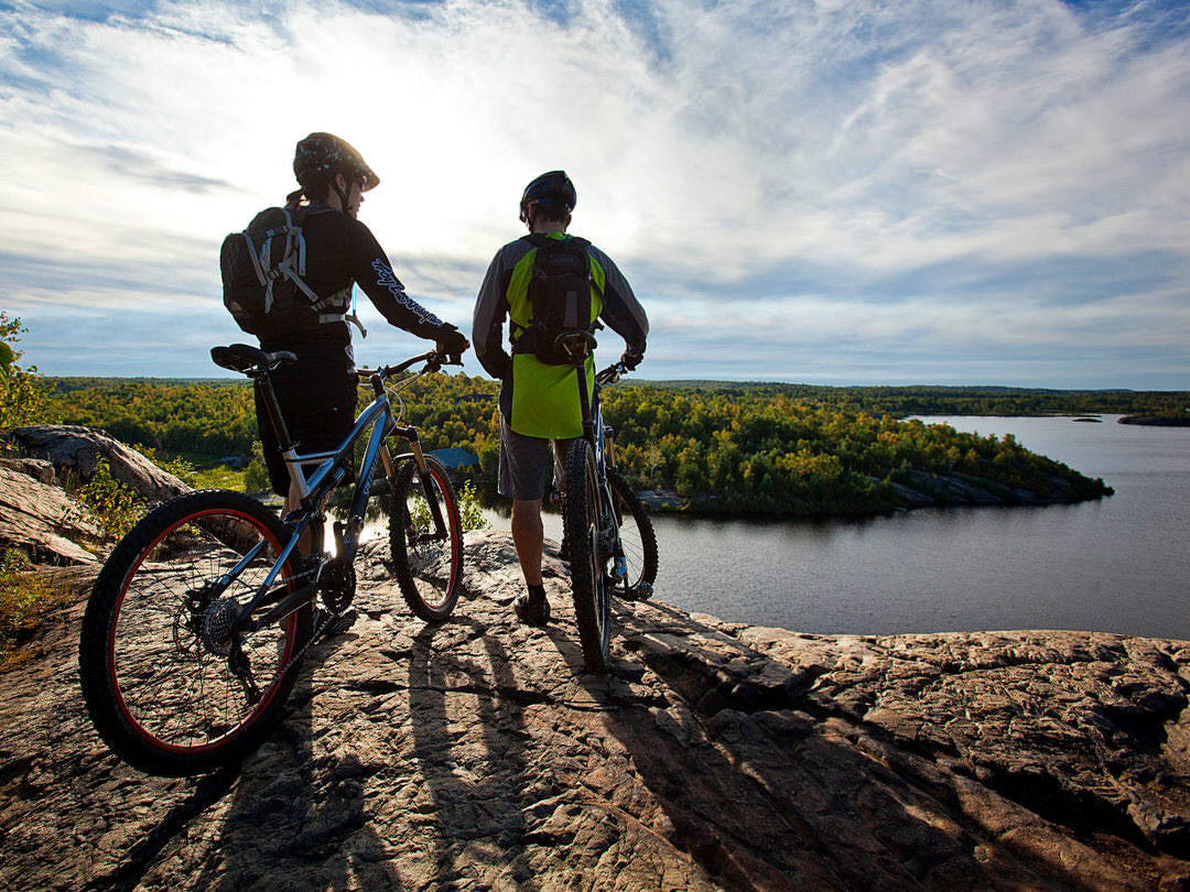 Two cyclists stand on the edge of a rock overlooking a scenic bay