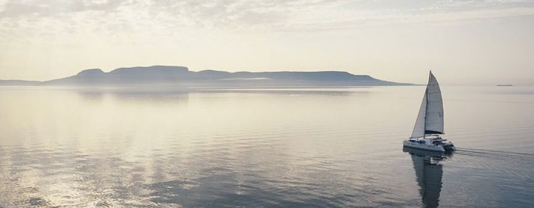 Reflections on water of a catamaran sailing towards hills in the distance as the sky is illuminated a purple gray