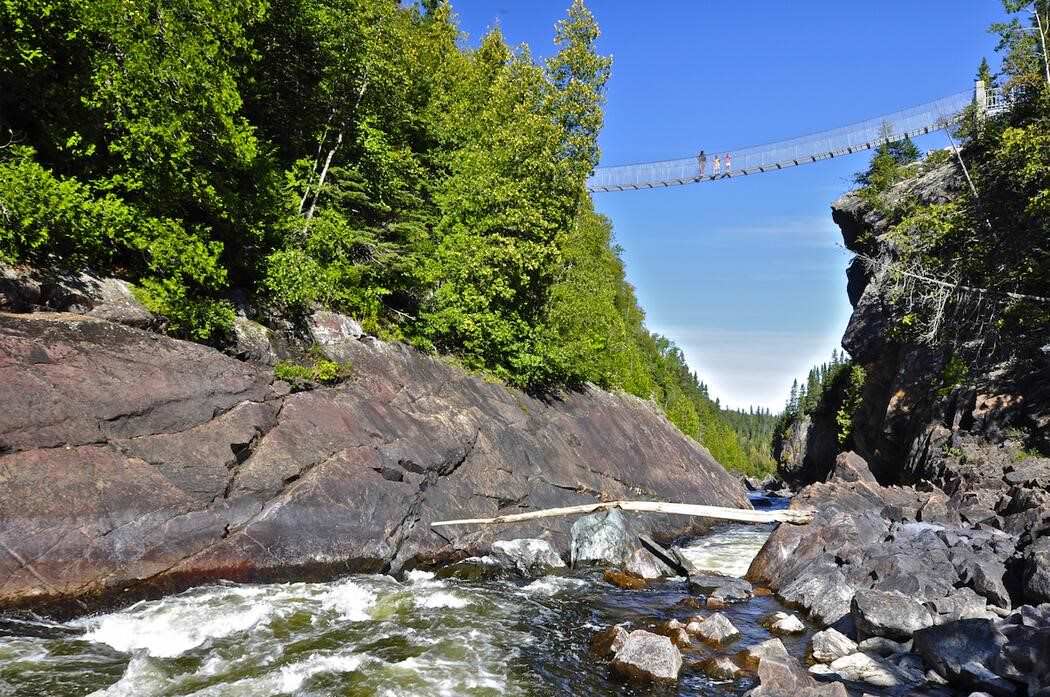3 personas emergen de un sendero para caminar en un bosque hacia un puente colgante, con vista a las rocas y un arroyo.
