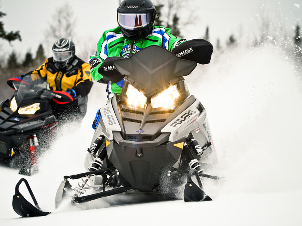 Deux personnes conduisent des motoneiges qui pulvérisent de la neige derrière elles.