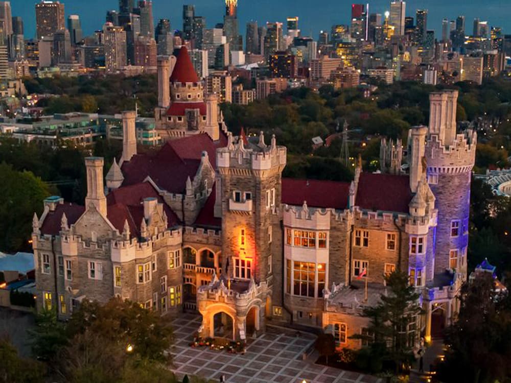 Aerial view of Casa Loma lit up at dusk with the city skyline behind.
