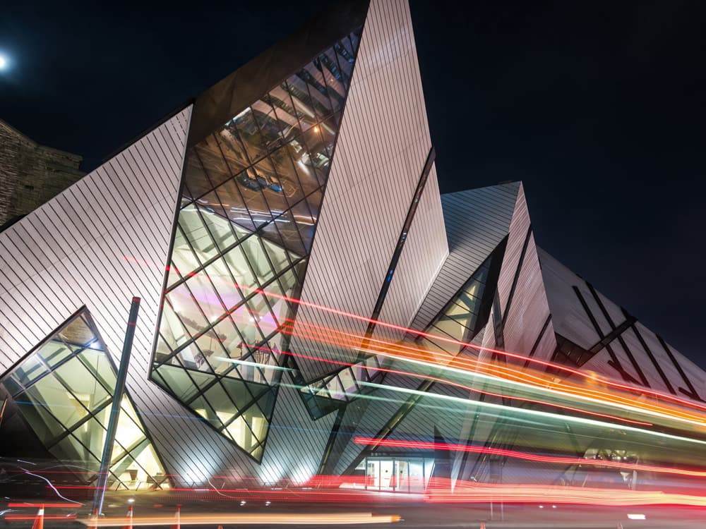 Geometrisch geformtes, modernes Gebäude hinter stark befahrener Straße bei Nacht