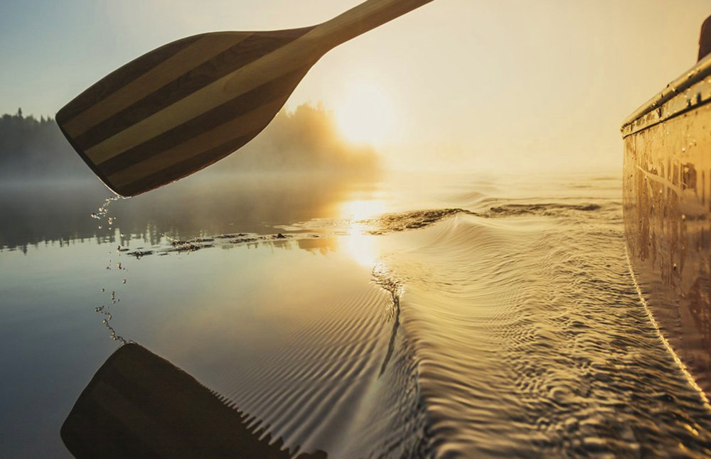 A canoe paddle gently rows through water off into a sunset.