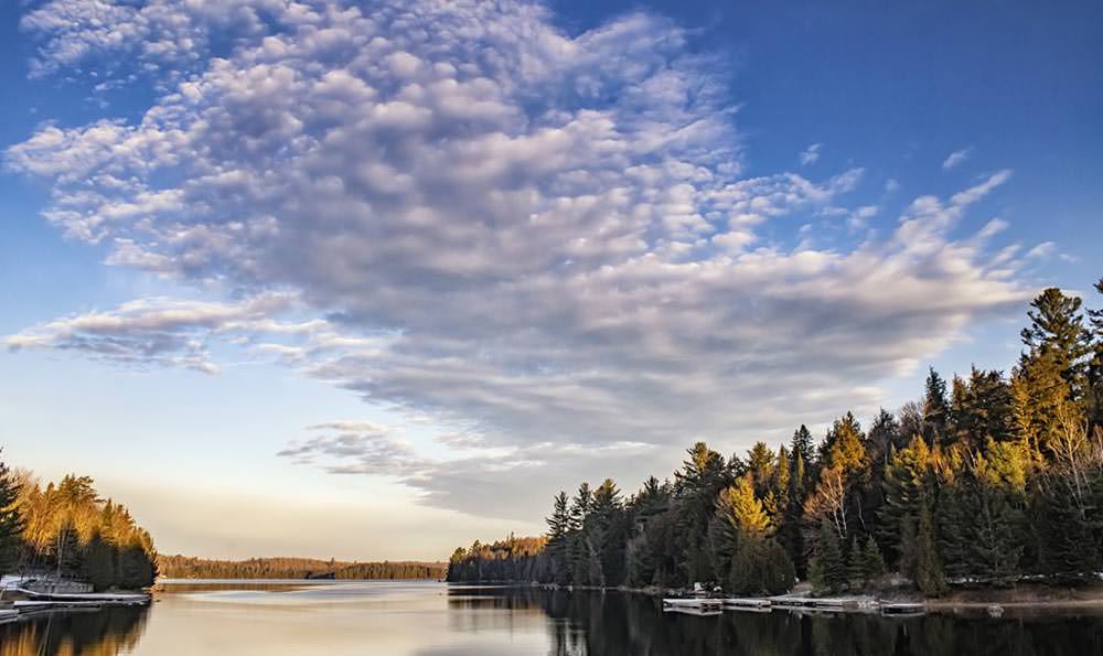 Ein blauer Himmel mit flauschigen Wolken spiegelte sich auf einem See, umgeben von einem Land voller Bäume.