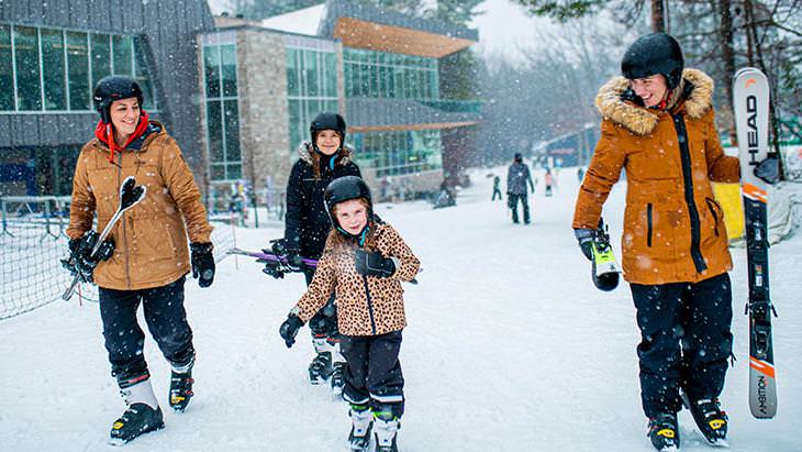 笑顔の大人 2 人と子供 2 人が、スキー板を持ってスキー ブーツを履いて雪の上を歩いています。