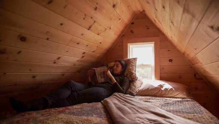 Eine junge Frau liegt auf einem Bett und liest ein Buch in einem gemütlichen holzgetäfelten Dachboden.