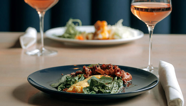 Dos platos de comida y dos copas de vino rosado se sientan en una mesa.